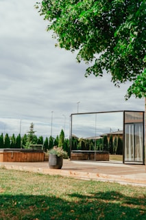 A serene outdoor scene featuring a wooden deck with a modern, glass-walled structure, nearby potted plants, and well-maintained green grass. Tall evergreen shrubs and trees create a natural boundary in the background, under a sky filled with light, fluffy clouds.