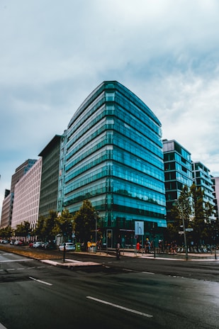 A modern urban setting featuring a large, multi-story glass office building. The architecture is sleek with reflective windows, situated on a city street with cars and pedestrians. Trees line the sidewalk, and the atmosphere suggests a busy, active area.