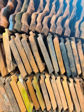 A stack of weathered clay roof tiles arranged in a symmetrical, overlapping pattern. The tiles have a rustic appearance with varying shades of orange, brown, and hints of green moss on some of them. They display a textured surface with visible aging and wear.