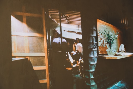 A dimly lit interior space with a group of people gathering near a wooden entrance. The room has a rustic design with wooden panels and a shelf with a vase containing green plants. The atmosphere is cozy, with warm lighting casting soft shadows.