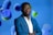 A man in a blue suit stands confidently with arms crossed against a vibrant background featuring abstract, colorful spheres in shades of blue, green, and purple. He is wearing a white shirt under the suit.