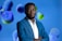 A man in a blue suit stands confidently with arms crossed against a vibrant background featuring abstract, colorful spheres in shades of blue, green, and purple. He is wearing a white shirt under the suit.