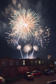 Bright fireworks light up the night sky, with large bursts of orange, white, and gold colors. The display takes place over a cityscape, featuring buildings with dark windows and parked cars along the street. The scene captures the contrast between the lively fireworks and the quiet urban environment below.