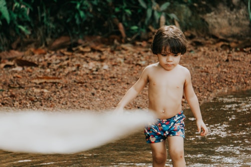 A young child with short brown hair is walking in shallow water near a sandy and rocky shore, wearing colorful, patterned swim shorts. Dense, green foliage can be seen in the background, adding to the natural setting.
