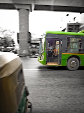 A green public bus is moving on a city street with a driver visible inside. In the foreground, part of an auto rickshaw is seen, adding to the urban traffic scene. The background includes an overhead metro track and blurred cityscape elements, suggesting motion and a bustling city environment.