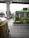 A green public bus is moving on a city street with a driver visible inside. In the foreground, part of an auto rickshaw is seen, adding to the urban traffic scene. The background includes an overhead metro track and blurred cityscape elements, suggesting motion and a bustling city environment.
