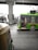 A green public bus is moving on a city street with a driver visible inside. In the foreground, part of an auto rickshaw is seen, adding to the urban traffic scene. The background includes an overhead metro track and blurred cityscape elements, suggesting motion and a bustling city environment.