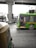 A green public bus is moving on a city street with a driver visible inside. In the foreground, part of an auto rickshaw is seen, adding to the urban traffic scene. The background includes an overhead metro track and blurred cityscape elements, suggesting motion and a bustling city environment.