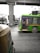 A green public bus is moving on a city street with a driver visible inside. In the foreground, part of an auto rickshaw is seen, adding to the urban traffic scene. The background includes an overhead metro track and blurred cityscape elements, suggesting motion and a bustling city environment.