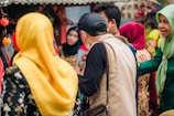 A group of people engaging in conversation. Many are dressed in colorful traditional attire, including headscarves of various vibrant colors such as yellow and red. The background features some festive decorations and a rustic structure.