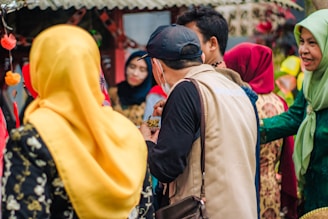 A group of people engaging in conversation. Many are dressed in colorful traditional attire, including headscarves of various vibrant colors such as yellow and red. The background features some festive decorations and a rustic structure.