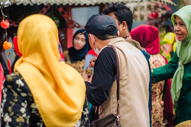 A group of people engaging in conversation. Many are dressed in colorful traditional attire, including headscarves of various vibrant colors such as yellow and red. The background features some festive decorations and a rustic structure.