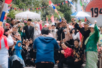 A large group of people are gathered outdoors, celebrating and posing cheerfully for a photo. Many are dressed in traditional attire, and some are wearing festive costumes. Brightly colored flags and banners add to the lively atmosphere, with trees visible in the background.