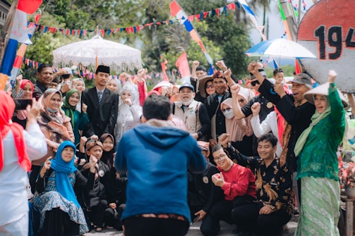 A large group of people are gathered outdoors, celebrating and posing cheerfully for a photo. Many are dressed in traditional attire, and some are wearing festive costumes. Brightly colored flags and banners add to the lively atmosphere, with trees visible in the background.