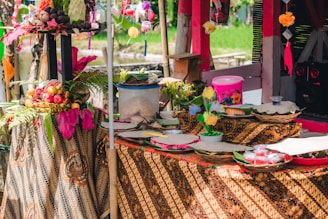 A vibrant and colorful setup featuring a table adorned with traditional batik cloth. Various decorative items are displayed, including baskets of fruits, ornate flowers, and small containers. The setting appears festive with bright yellow and pink accents along with touches of greenery. Sunlight filters through, adding a natural, warm ambiance.