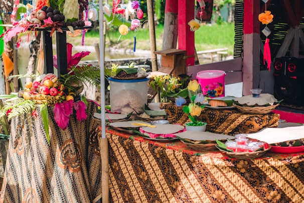A vibrant and colorful setup featuring a table adorned with traditional batik cloth. Various decorative items are displayed, including baskets of fruits, ornate flowers, and small containers. The setting appears festive with bright yellow and pink accents along with touches of greenery. Sunlight filters through, adding a natural, warm ambiance.