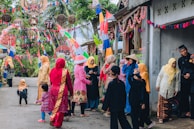 A group of people dressed in colorful traditional outfits, gathered on a street decorated with vibrant flags and banners. The scene is festive, with women wearing headscarves and children playing nearby. The street is lined with small houses and lush greenery in the background.