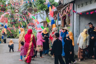 A group of people dressed in colorful traditional outfits, gathered on a street decorated with vibrant flags and banners. The scene is festive, with women wearing headscarves and children playing nearby. The street is lined with small houses and lush greenery in the background.