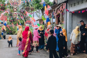 A group of people dressed in colorful traditional outfits, gathered on a street decorated with vibrant flags and banners. The scene is festive, with women wearing headscarves and children playing nearby. The street is lined with small houses and lush greenery in the background.