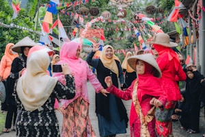 A group of people dressed in bright, colorful traditional clothing are gathered outdoors. Some are wearing hats and hijabs, and they appear to be celebrating or participating in a festive event. Bunting and flags are strung overhead, adding to the lively atmosphere. The setting is a vibrant street surrounded by greenery.