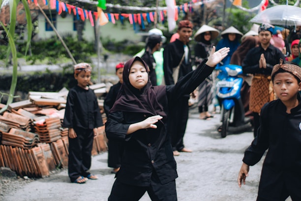 A group of individuals dressed in traditional clothing are gathered outdoors. A young person in the foreground strikes a pose with arms extended, suggesting a dance or martial art movement. Others in the background are wearing hats and various attire, with some appearing to observe the performance. Brightly colored flags hang overhead, and there is a blue scooter parked nearby.