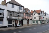 A picturesque street scene featuring a row of historic timber-framed buildings. The architecture showcases a blend of black and white wooden beams with white and pastel-colored walls. The structures house various shops, including one labeled 'White Rose Gallery.' Pedestrians are walking along the sidewalk.