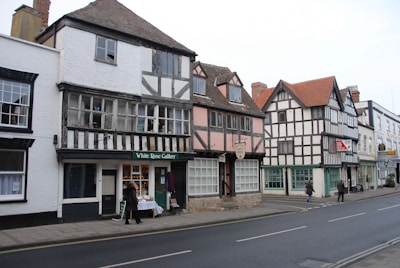 A picturesque street scene featuring a row of historic timber-framed buildings. The architecture showcases a blend of black and white wooden beams with white and pastel-colored walls. The structures house various shops, including one labeled 'White Rose Gallery.' Pedestrians are walking along the sidewalk.