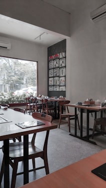 A cozy, modern restaurant interior with wooden tables and chairs arranged neatly. The walls are adorned with a grid of artistic panels, and a large window offers a view of greenery and the street outside. Natural lighting creates a serene and inviting ambiance.