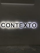 Illuminated letters spelling out the word 'CONTEXTO' are mounted on a wall. The black letters are backlit, creating a halo effect against the light gray wall.