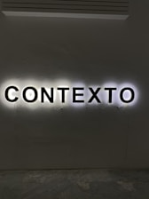 Illuminated letters spelling out the word 'CONTEXTO' are mounted on a wall. The black letters are backlit, creating a halo effect against the light gray wall.
