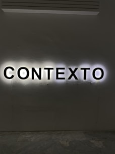 Illuminated letters spelling out the word 'CONTEXTO' are mounted on a wall. The black letters are backlit, creating a halo effect against the light gray wall.