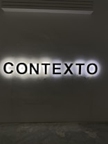 Illuminated letters spelling out the word 'CONTEXTO' are mounted on a wall. The black letters are backlit, creating a halo effect against the light gray wall.