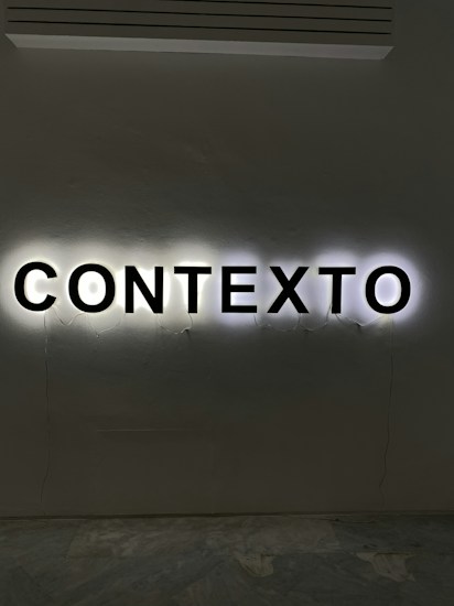 Illuminated letters spelling out the word 'CONTEXTO' are mounted on a wall. The black letters are backlit, creating a halo effect against the light gray wall.