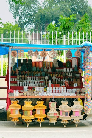A street stall displays a variety of decorative items and souvenirs. Intricate statues, sculptures, and hanging crafts are neatly arranged on shelves. The setup is colorful, featuring pottery, religious symbols, and other handmade artifacts. The stall is covered by a blue tarp, and greenery can be seen in the background.