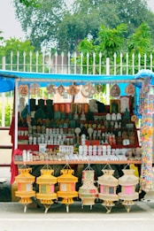 A street stall displays a variety of decorative items and souvenirs. Intricate statues, sculptures, and hanging crafts are neatly arranged on shelves. The setup is colorful, featuring pottery, religious symbols, and other handmade artifacts. The stall is covered by a blue tarp, and greenery can be seen in the background.