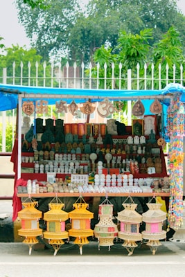 A street stall displays a variety of decorative items and souvenirs. Intricate statues, sculptures, and hanging crafts are neatly arranged on shelves. The setup is colorful, featuring pottery, religious symbols, and other handmade artifacts. The stall is covered by a blue tarp, and greenery can be seen in the background.