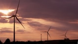 Tall wind turbines stand against a backdrop of a dramatic sunset sky, with sunlight piercing through scattered clouds. The silhouetted turbines create a serene yet striking image, emphasizing both the grandeur of the sky and the elegance of renewable energy structures.