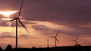 Tall wind turbines stand against a backdrop of a dramatic sunset sky, with sunlight piercing through scattered clouds. The silhouetted turbines create a serene yet striking image, emphasizing both the grandeur of the sky and the elegance of renewable energy structures.