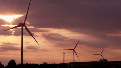 Tall wind turbines stand against a backdrop of a dramatic sunset sky, with sunlight piercing through scattered clouds. The silhouetted turbines create a serene yet striking image, emphasizing both the grandeur of the sky and the elegance of renewable energy structures.