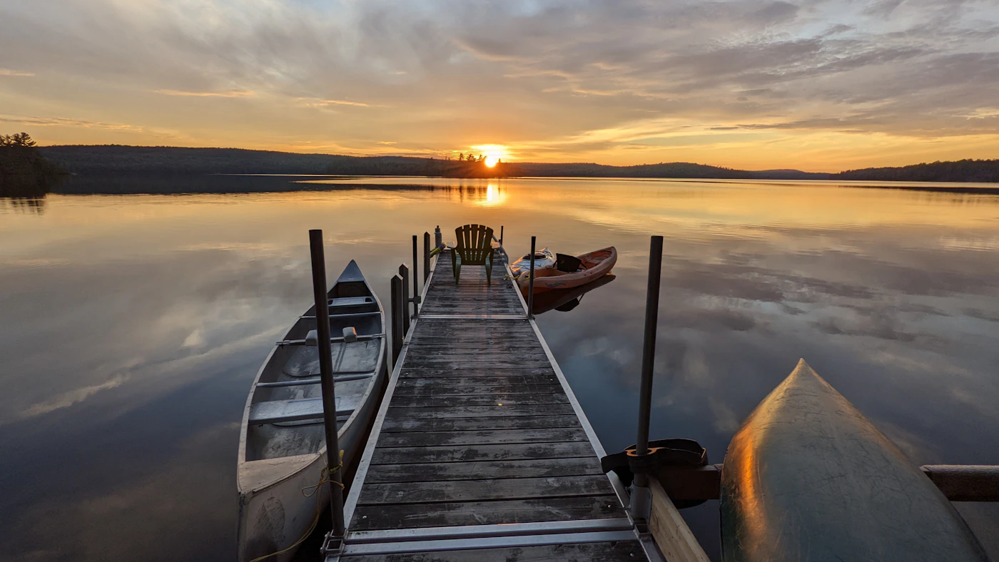 Wooden dock extending over a calm Ontario lake at sunset with two canoes tied alongside, a Muskoka chair, and the sun setting in warm orange above the forested horizon