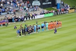 A soccer match scene at a stadium shows two teams and referees standing in formation on the field. One team wears black uniforms while the other wears orange. Several people, including photographers and children, stand in line or kneel in front of them. The background shows a crowd of spectators seated in the stands, banners from sponsors, and a large Stella Artois sign.