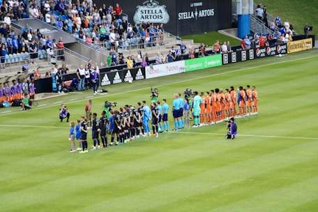 A soccer match scene at a stadium shows two teams and referees standing in formation on the field. One team wears black uniforms while the other wears orange. Several people, including photographers and children, stand in line or kneel in front of them. The background shows a crowd of spectators seated in the stands, banners from sponsors, and a large Stella Artois sign.
