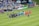 A soccer match scene at a stadium shows two teams and referees standing in formation on the field. One team wears black uniforms while the other wears orange. Several people, including photographers and children, stand in line or kneel in front of them. The background shows a crowd of spectators seated in the stands, banners from sponsors, and a large Stella Artois sign.
