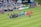 A soccer match scene at a stadium shows two teams and referees standing in formation on the field. One team wears black uniforms while the other wears orange. Several people, including photographers and children, stand in line or kneel in front of them. The background shows a crowd of spectators seated in the stands, banners from sponsors, and a large Stella Artois sign.