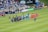 A soccer match scene at a stadium shows two teams and referees standing in formation on the field. One team wears black uniforms while the other wears orange. Several people, including photographers and children, stand in line or kneel in front of them. The background shows a crowd of spectators seated in the stands, banners from sponsors, and a large Stella Artois sign.