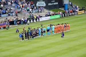 A soccer match scene at a stadium shows two teams and referees standing in formation on the field. One team wears black uniforms while the other wears orange. Several people, including photographers and children, stand in line or kneel in front of them. The background shows a crowd of spectators seated in the stands, banners from sponsors, and a large Stella Artois sign.