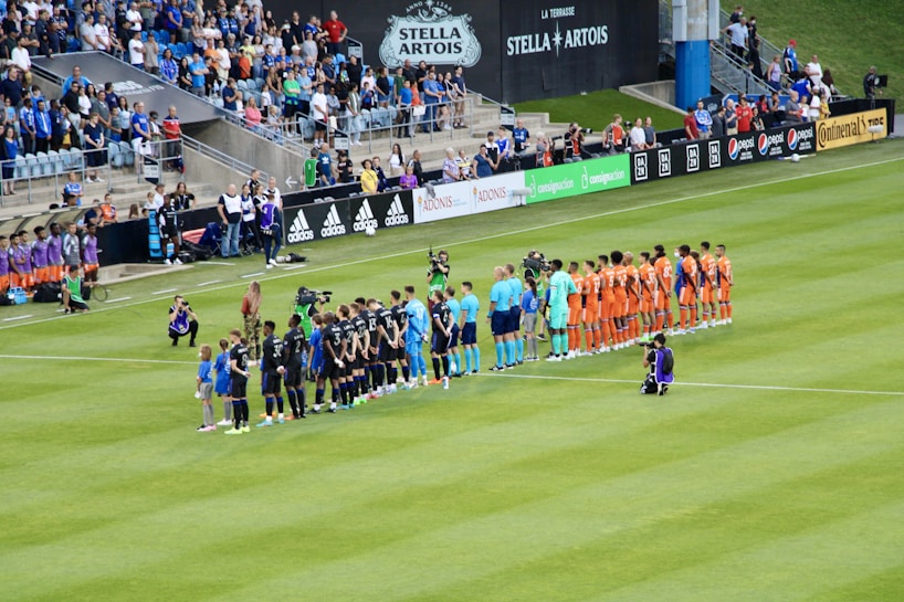 A soccer match scene at a stadium shows two teams and referees standing in formation on the field. One team wears black uniforms while the other wears orange. Several people, including photographers and children, stand in line or kneel in front of them. The background shows a crowd of spectators seated in the stands, banners from sponsors, and a large Stella Artois sign.