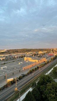 An aerial view of an urban area featuring a complex of industrial buildings and a railway line running alongside a road. The sky is cloudy, and the sunlit rooftops and surroundings suggest either early morning or late afternoon. Vegetation and trees can be seen beside the railway tracks.