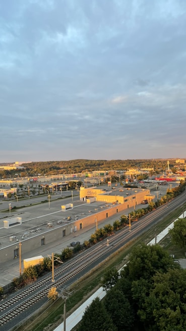 An aerial view of an urban area featuring a complex of industrial buildings and a railway line running alongside a road. The sky is cloudy, and the sunlit rooftops and surroundings suggest either early morning or late afternoon. Vegetation and trees can be seen beside the railway tracks.