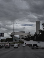 A busy road with multiple cars heading towards a prominent structure that includes a large archway labeled 'Tecnológico de Monterrey'. The background shows a tall building with a distinct logo near the top, surrounded by trees and greenery. The sky above is overcast with thick clouds, contributing to a somewhat gloomy atmosphere.